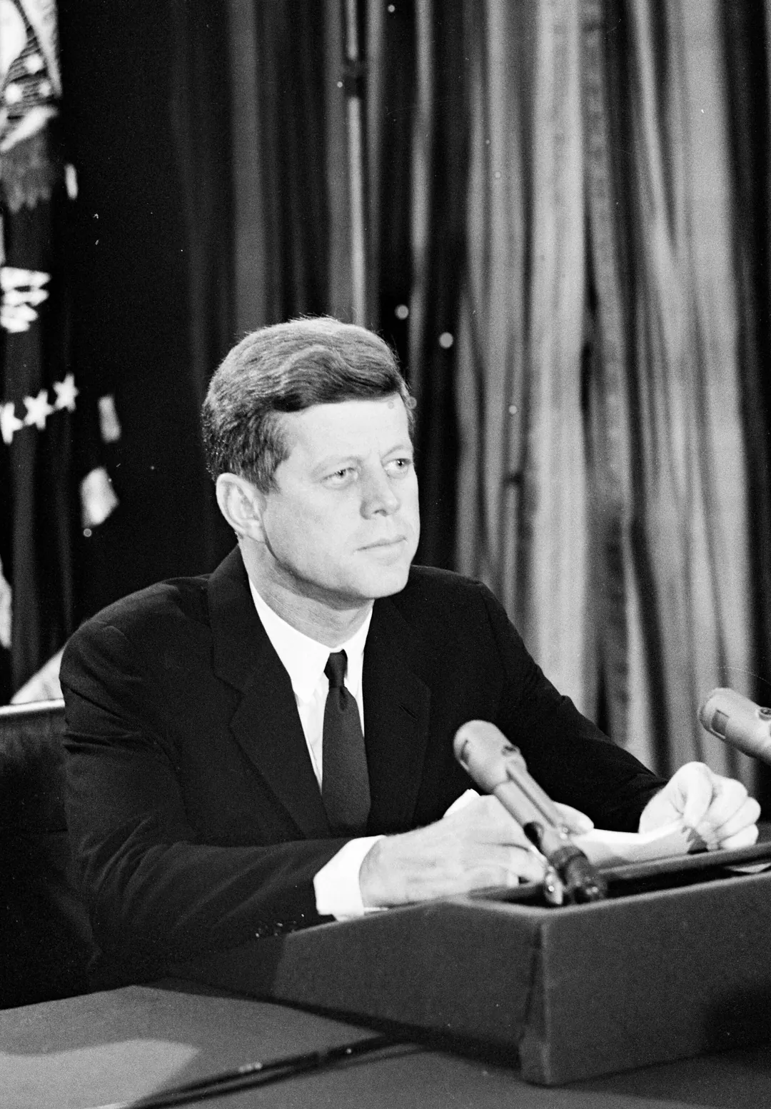 Black-and-white photo: Kennedy at a desk with microphones and the pages of the speech during the address to the nation, October 22, 1962